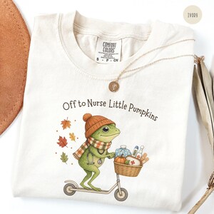 May include: An ivory t-shirt featuring a graphic of a green frog wearing an orange knit hat and plaid scarf, riding a scooter. The frog has a stethoscope and a basket containing a pumpkin and medical supplies. Text reads: Off to Nurse Little Pumpkins.