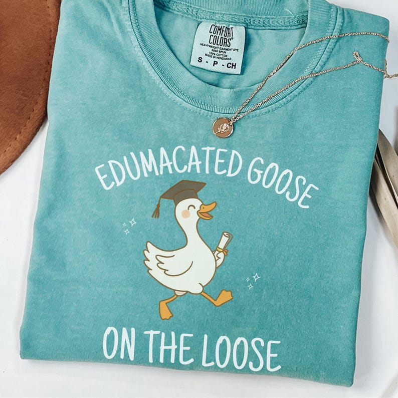 K&ouml;nnte beinhalten: Ein t&uuml;rkisfarbenes T-Shirt mit einer wei&szlig;en Grafik einer Gans, die einen Abschluss-Hut tr&auml;gt und ein Diplom h&auml;lt. Der Text "Edumacated Goose on the Loose" ist auf das T-Shirt gedruckt.
