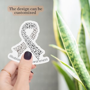 May include: A white sticker in the shape of a ribbon, composed of black ribbon icons, with the words "Glioblastoma Awareness." The text "The design can be customized" is above the sticker. The sticker is held by a hand with a dark purple nail.
