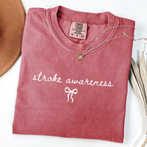 May include: A dusty rose Comfort Colors t-shirt with the words "stroke awareness" embroidered in white, along with a bow graphic. A silver necklace with a small pendant rests on the shirt. A brown hat is partially visible in the upper left corner.