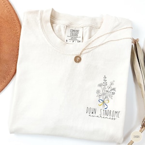Op de afbeelding: Een ivoorkleurig t-shirt met een zwarte lijntekening van een boeket wilde bloemen vastgebonden met een blauw en geel lint. Onder het boeket staat de tekst Down Syndrome awareness. Het t-shirt heeft een Comfort Colors label in de binnenhals.