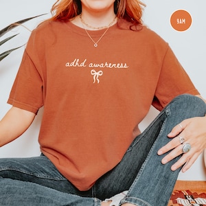 May include: A rust-colored t-shirt with the words "adhd awareness" in cursive script and a bow graphic. The shirt is a short-sleeved, crew neck style. The model is wearing blue jeans and jewelry.