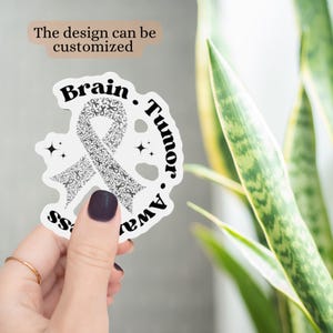 May include: A white sticker with a brain tumor awareness ribbon design. The ribbon is made of small, repeating shapes. The words "Brain Tumor Awareness" are printed around the ribbon in a circular pattern. The text "The design can be customized" is also present.