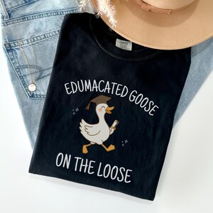 K&ouml;nnte beinhalten: Schwarzes T-Shirt mit einer wei&szlig;en Grafik einer Gans, die einen Abschluss-Hut tr&auml;gt und ein Diplom h&auml;lt. Der Text "Edumacated Goose on the Loose" ist auf das T-Shirt gedruckt.