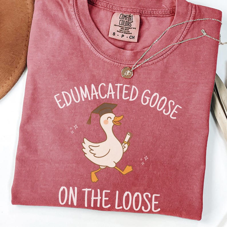 K&ouml;nnte beinhalten: Ein rotes T-Shirt mit dem Text "Edumacated Goose on the Loose" und einer Cartoon-Illustration einer Gans, die einen Abschluss-Hut tr&auml;gt und ein Diplom h&auml;lt.