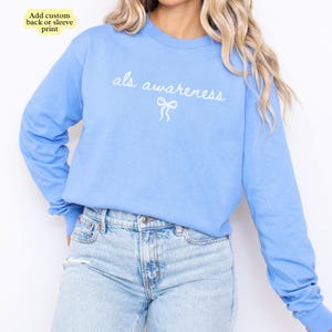 May include: A light blue long-sleeve sweatshirt with the words "als awareness" in white script and a bow graphic. The sweatshirt is paired with light wash denim jeans. The text "Add custom back or sleeve print" is in the top left corner.