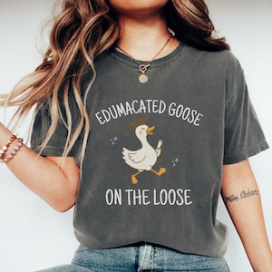 K&ouml;nnte beinhalten: Ein dunkelgraues T-Shirt mit einer wei&szlig;en Grafik einer Gans, die einen Abschluss-Hut tr&auml;gt und ein Diplom h&auml;lt. Der Text "Edumacated Goose on the Loose" ist auf das T-Shirt gedruckt.