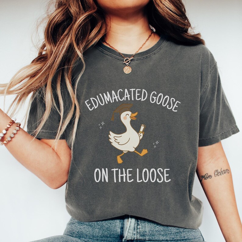 K&ouml;nnte beinhalten: Ein dunkelgraues T-Shirt mit einer wei&szlig;en Grafik einer Gans, die einen Abschluss-Hut tr&auml;gt und ein Diplom h&auml;lt. Der Text "Edumacated Goose on the Loose" ist auf das T-Shirt gedruckt.