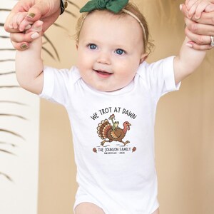 May include: A white short-sleeved baby bodysuit featuring a graphic of a turkey with a frog on its back. The text reads WE TROT AT DAWN, THE JOHNSON FAMILY, and NASHVILLE 2026, accompanied by small orange leaf illustrations on either side of the text.