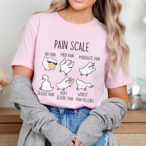 Pu&ograve; includere: Una maglietta rosa con una grafica di scala del dolore che presenta una serie di oche dei cartoni animati con diverse espressioni. Il testo recita "Pain Scale", "No Pain", "Mild Pain", "Moderate Pain", "Severe Pain", "Very Severe Pain", e "Worst Pain Possible".