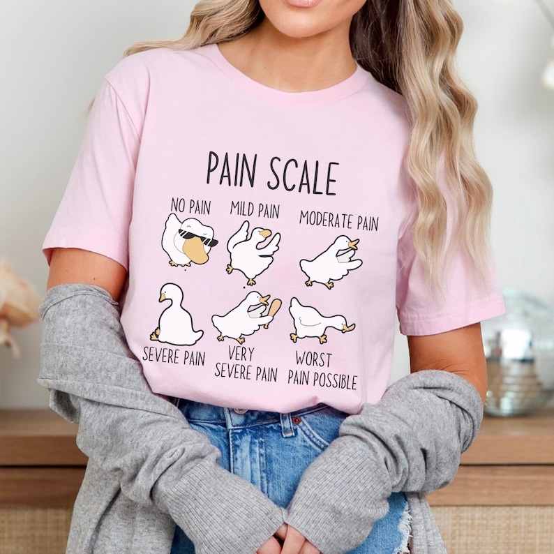 Pu&ograve; includere: Una maglietta rosa con una grafica di scala del dolore che presenta una serie di oche dei cartoni animati con diverse espressioni. Il testo recita "Pain Scale", "No Pain", "Mild Pain", "Moderate Pain", "Severe Pain", "Very Severe Pain", e "Worst Pain Possible".