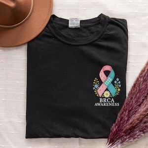 May include: Black t-shirt featuring a pink and blue ribbon design with the words "BRCA AWARENESS" in white. The ribbon is surrounded by small flowers. A brown hat is visible in the upper left corner.
