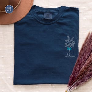 May include: A navy blue t-shirt with a small floral design and the text "ALS awareness" in white. The shirt has a crew neck and short sleeves. A brown hat is visible in the upper left corner.