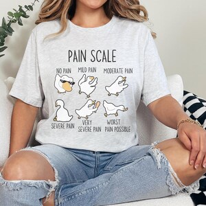 Pu&ograve; includere: Una maglietta grigia con una grafica di scala del dolore che presenta una serie di oche dei cartoni animati con diverse espressioni. Il testo recita "Pain Scale" e include descrizioni come "No Pain", "Mild Pain", "Moderate Pain", "Severe Pain", "Very Severe Pain" e "Worst Pain Possible".