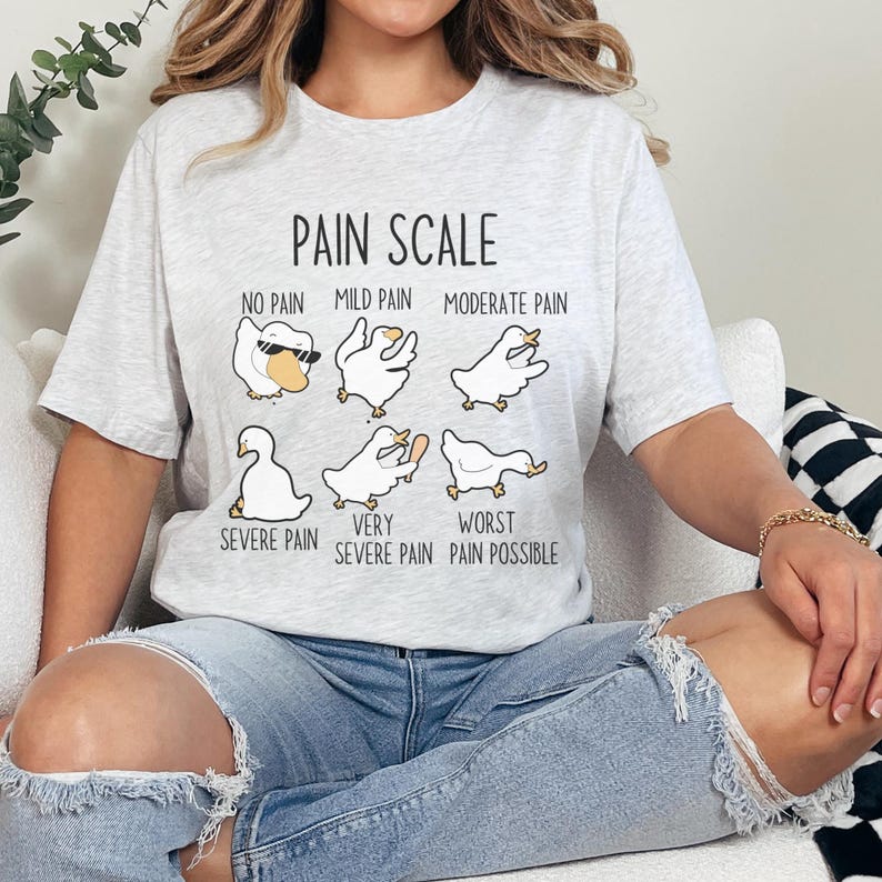 Pu&ograve; includere: Una maglietta grigia con una grafica di scala del dolore che presenta una serie di oche dei cartoni animati con diverse espressioni. Il testo recita "Pain Scale" e include descrizioni come "No Pain", "Mild Pain", "Moderate Pain", "Severe Pain", "Very Severe Pain" e "Worst Pain Possible".