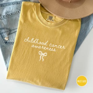 May include: A mustard yellow t-shirt featuring the white text "childhood cancer awareness" and a small white ribbon graphic. The shirt has a crew neckline and a Comfort Colors label. A tan hat and blue denim fabric are visible in the background.
