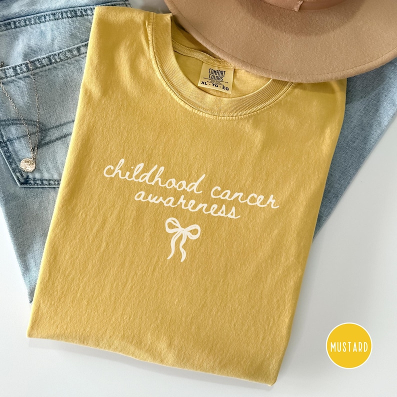 May include: A mustard yellow t-shirt featuring the white text "childhood cancer awareness" and a small white ribbon graphic. The shirt has a crew neckline and a Comfort Colors label. A tan hat and blue denim fabric are visible in the background.
