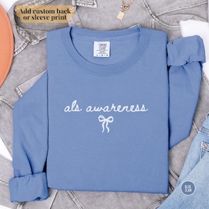 May include: A light blue long-sleeve shirt with the words "als awareness" and a bow graphic in white. The shirt is folded and displayed on a denim background. The shirt is labeled "Comfort Colors".