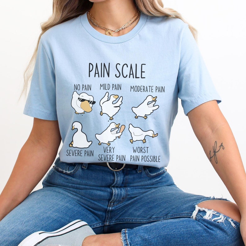 Pu&ograve; includere: Una maglietta blu chiaro con una grafica di una scala del dolore che utilizza anatre dei cartoni animati. Il testo sulla maglietta recita "Pain Scale", "No Pain", "Mild Pain", "Moderate Pain", "Severe Pain", "Very Severe Pain", e "Worst Pain Possible".