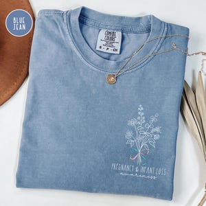 May include: A blue jean colored short-sleeve t-shirt with a crew neckline. The shirt features a white line art graphic of a wildflower bouquet tied with a pink and blue ribbon, accompanied by the text Pregnancy & Infant Loss Awareness.