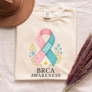May include: Cream-colored t-shirt featuring a pink and teal awareness ribbon design with floral accents and the text "BRCA AWARENESS". The ribbon has a floral and gingham pattern. The shirt is a Comfort Colors XL size.