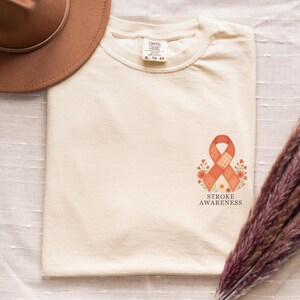May include: Cream-colored t-shirt with a printed orange ribbon and floral design, with the words "STROKE AWARENESS" below. A brown hat is visible in the upper left corner. The shirt is a Comfort Colors XL-TG-EG.
