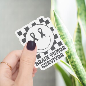 May include: A die-cut sticker featuring a white smiley face with gray ribbon-shaped eyes, set against a black and white checkered background. The text "BRAIN TUMOR SURVIVOR" is printed in a bold, dark gray font at the bottom of the sticker.
