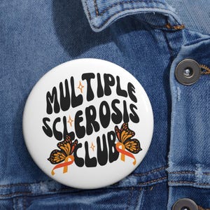 May include: A white button with the text "MULTIPLE SCLEROSIS CLUB" in black, stylized font. Two orange and black butterflies and orange ribbons are featured below the text. The button is set against a blue denim background.