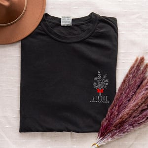 May include: Black t-shirt with a floral design and the words "Stroke awareness" in white. The design includes a red bow. A brown hat is visible in the upper left corner, and a bundle of dried flowers is in the lower right.