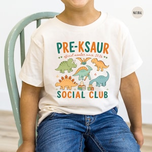 May include: A natural-colored t-shirt featuring a graphic print with the text PRE-KSAUR SOCIAL CLUB and official member since 2026. The design includes colorful illustrations of various dinosaurs, a stack of books, and alphabet blocks.