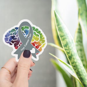 May include: A sticker featuring a gray awareness ribbon with the text "Your Custom Text" and a white heart. The ribbon is superimposed over a colorful brain design with watercolor effects. The sticker is held by a hand.