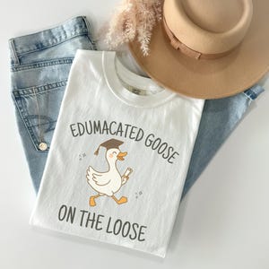 K&ouml;nnte beinhalten: Wei&szlig;es T-Shirt mit einer Cartoon-Illustration einer Gans, die einen Abschluss-Hut tr&auml;gt und ein Diplom h&auml;lt. Der Text "Edumacated Goose on the Loose" ist auf das Shirt gedruckt.