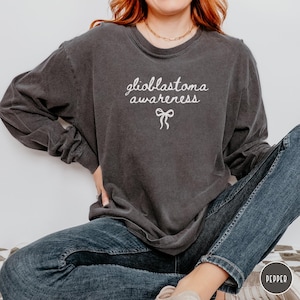 May include: Dark gray long-sleeve shirt with the words "glioblastoma awareness" in white script, and a white bow graphic. The shirt is made of a soft material and is paired with blue jeans. The shirt is a casual wear item.