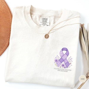May include: Ivory-colored t-shirt with a purple awareness ribbon design, surrounded by flowers, and the text "Ulcerative Colitis Awareness." A gold necklace with a pendant rests on the shirt. The shirt is labeled "Comfort Colors."
