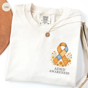 May include: Ivory-colored t-shirt with a crew neck and the text "ADHD AWARENESS". The shirt features a colorful ribbon design with flowers. A gold necklace with a pendant is draped across the shirt.