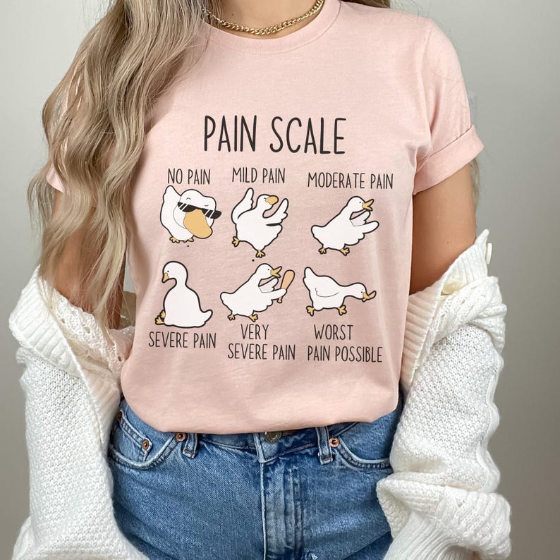Pu&ograve; includere: Una maglietta rosa con una grafica di scala del dolore che presenta una serie di oche dei cartoni animati con diverse espressioni. Il testo recita "Pain Scale", "No Pain", "Mild Pain", "Moderate Pain", "Severe Pain", "Very Severe Pain", e "Worst Pain Possible".
