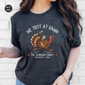 May include: A dark grey heather t-shirt featuring a graphic of a turkey with a frog riding on its back. The text reads WE TROT AT DAWN, THE JOHNSON FAMILY, and NASHVILLE 2026. The design includes two small orange leaves on either side of the text.