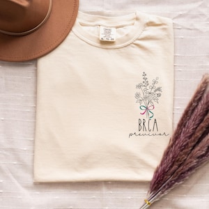May include: Cream-colored t-shirt with a floral bouquet design and the text "BRCA previvor" in black. The bouquet includes a pink, teal, and green ribbon. A brown hat is visible in the upper left corner.