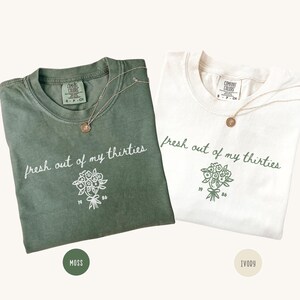 Può includere: Due magliette a girocollo in verde muschio e avorio, ciascuna con la scritta "fresh out of my thirties" in corsivo sopra un disegno al tratto di un bouquet di fiori con i numeri "19" e "86" su entrambi i lati dei gambi.