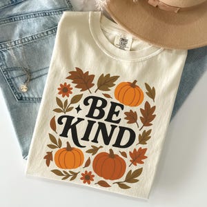 May include: Cream-colored t-shirt with a fall-themed graphic. The design features orange pumpkins, brown leaves, and the words "BE KIND" in black. A gold necklace, blue jeans, and a tan hat are also visible.