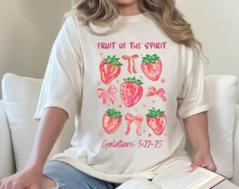 May include: Cream-colored t-shirt featuring a strawberry design with pink bows and the text "Fruit of the Spirit" and "Galatians 5:22-23" in pink. The strawberries have green leaves and pink accents.