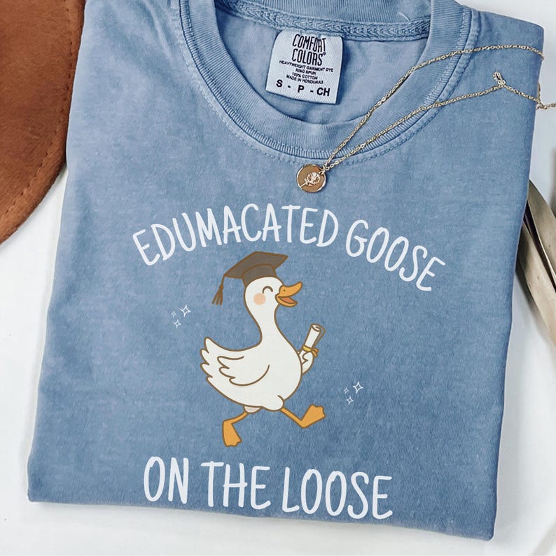 K&ouml;nnte beinhalten: Ein hellblaues T-Shirt mit einer Cartoon-Gans, die einen Abschluss-Hut tr&auml;gt und ein Diplom h&auml;lt. Der Text "Edumacated Goose on the Loose" ist auf das T-Shirt gedruckt.