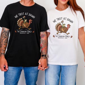 May include: A black t-shirt and a white t-shirt, each featuring a graphic of a turkey with a frog riding on its back. The text reads WE TROT AT DAWN, THE JOHNSON FAMILY, and NASHVILLE 2026. The turkey is brown with a red wattle and a green frog.