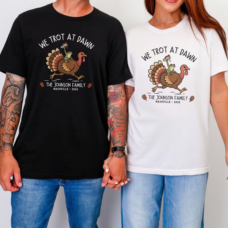 May include: A black t-shirt and a white t-shirt, each featuring a graphic of a turkey with a frog riding on its back. The text reads WE TROT AT DAWN, THE JOHNSON FAMILY, and NASHVILLE 2026. The turkey is brown with a red wattle and a green frog.
