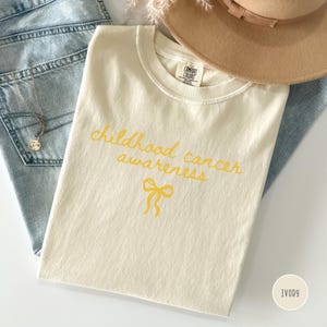 May include: An ivory t-shirt with the text "childhood cancer awareness" in yellow cursive script, featuring a yellow ribbon graphic below the text. The shirt has a Comfort Colors brand label at the inner neckline and is made of 100% cotton.