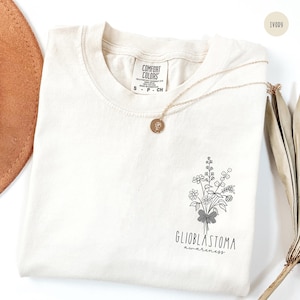 May include: Ivory-colored t-shirt with a floral graphic and the words "Glioblastoma awareness." A gold necklace with a round pendant rests on the shirt. The shirt has a Comfort Colors tag.