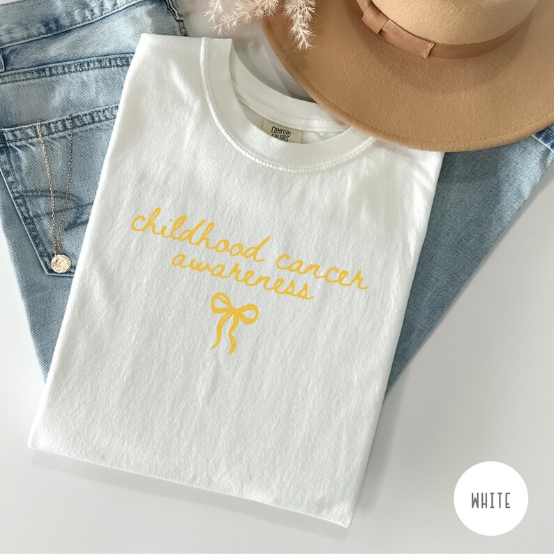 May include: A white t-shirt featuring the yellow cursive text childhood cancer awareness and a yellow ribbon graphic. The shirt is layered with light blue denim jeans, a tan wide-brimmed hat, and a silver chain necklace with a round pendant.