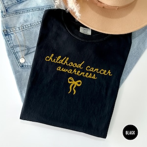 May include: A black t-shirt featuring the gold text "childhood cancer awareness" and a gold ribbon icon. The shirt has a Comfort Colors label at the inner neckline. The garment is displayed alongside light blue denim fabric and a tan hat.