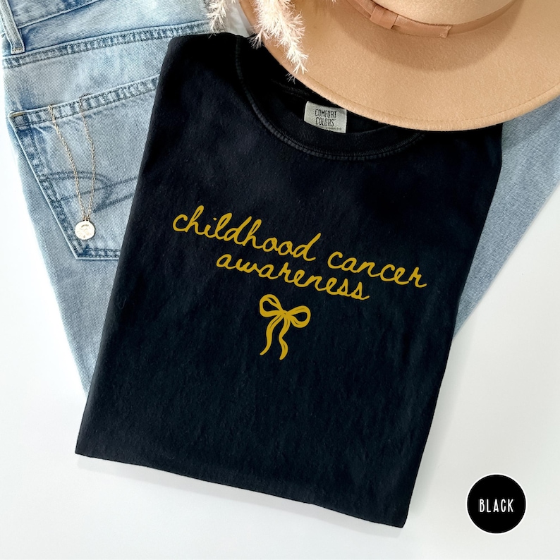 May include: A black t-shirt featuring the gold text "childhood cancer awareness" and a gold ribbon icon. The shirt has a Comfort Colors label at the inner neckline. The garment is displayed alongside light blue denim fabric and a tan hat.