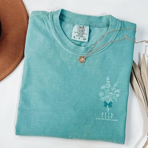 May include: A teal Comfort Colors t-shirt with a floral design and the words "PTSD awareness" printed on the front. A gold necklace with a pendant and a brown hat are also visible.